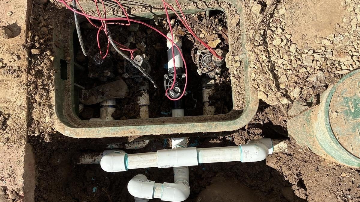 Open sprinkler valve box with the manifold and wiring exposed, and new PVC piping run below it.