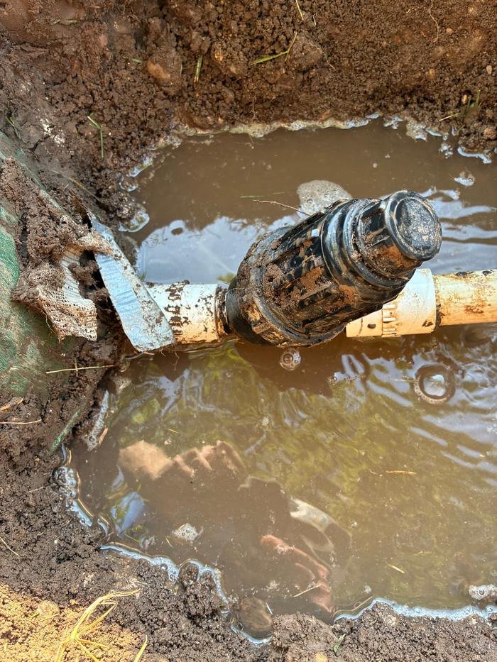 Sprinkler lateral leaking into a water-filled trench, its joint patched with cloth tape.